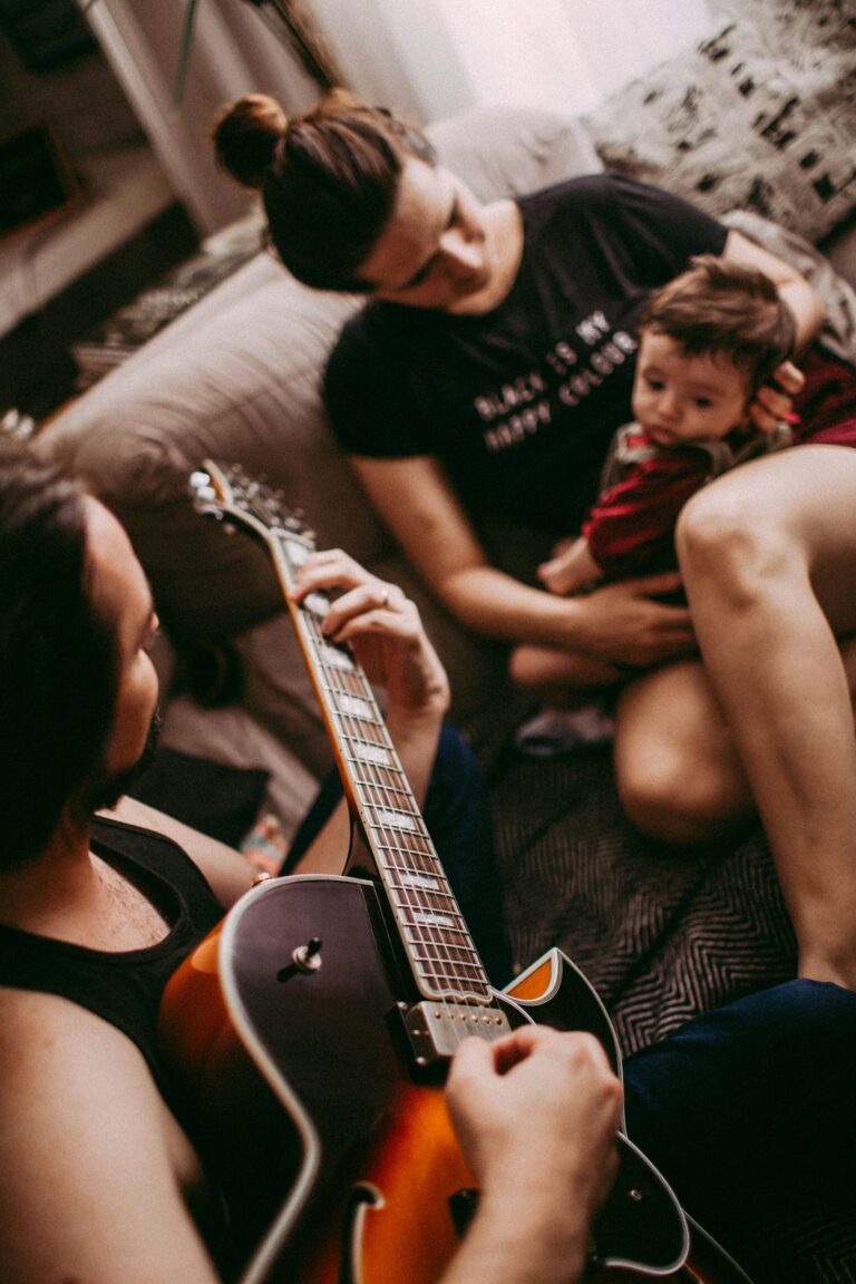 From above of crop unrecognizable ethnic musician playing guitar near woman embracing child on cozy sofa at home
